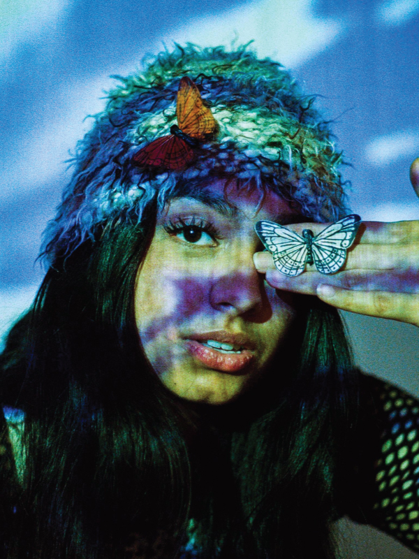 A woman wears a blue bucket hat and poses in a close up shot with a butterfly on her hand. Her face is illuminated by blue projections