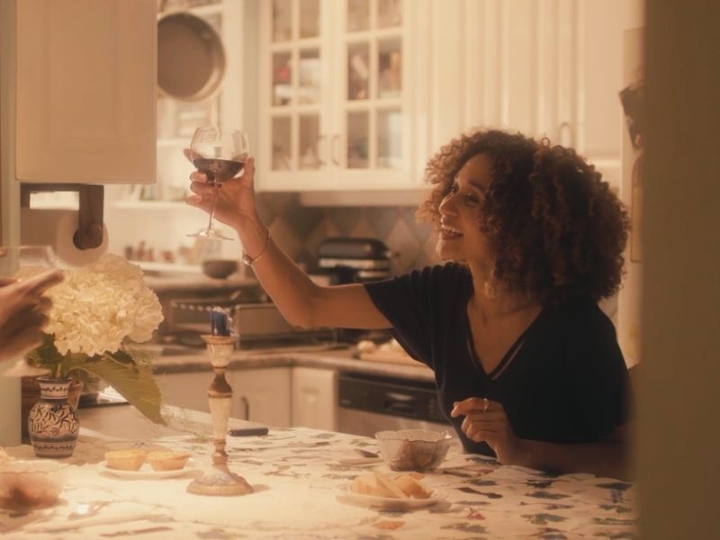 A young black woman sitting at a dinner table lifts a glass of red wine in a toast