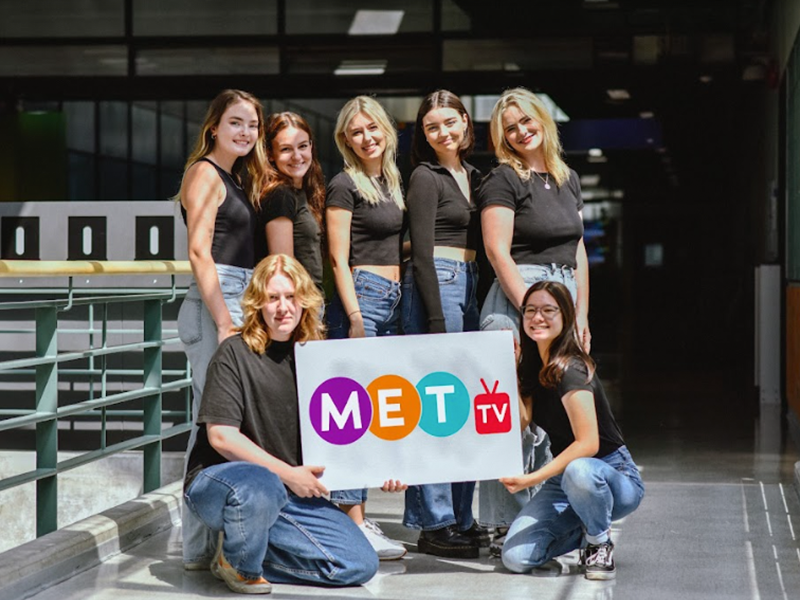 A group of women wearing black shirts and blue jeans stand together holding a sign that says Met-TV.