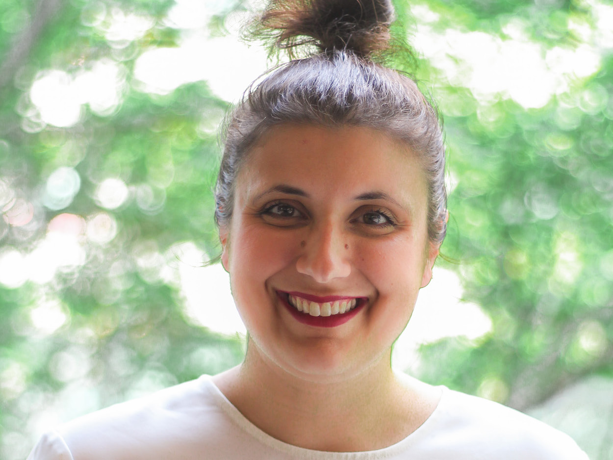 Director of Academic Planning and Student Affairs, Marie Crosta. Marie wears a white t-shirt and red lipstick and is smiling. Her hair is gathered in a bun.