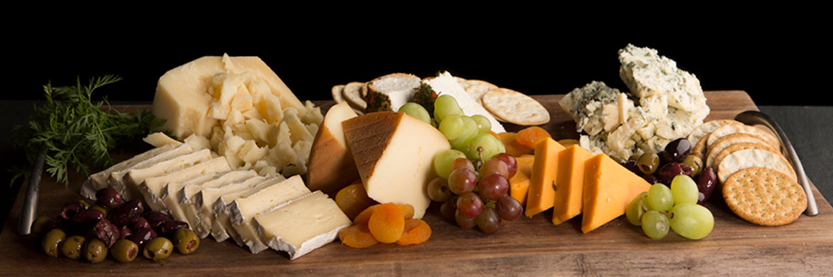 a charcuterie spread of cheese, crackers and fruit on a wooden board with a black background.