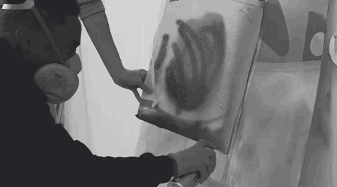 An over-the-shoulder image of Nuff spray-painting a wall. Someone is holding another piece of material over the surface as a mask. 