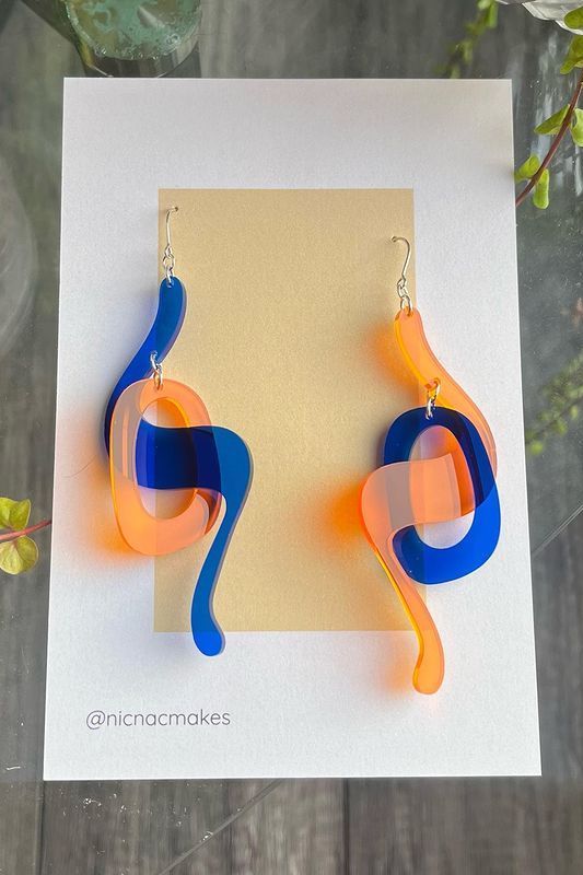 A pair or plastic earrings in nicnac-branded packaging. One earring has a transparent blue swish intersecting with a transparent orange loop. The other earring has opposite colors. The earring package rests on a wooden surface. 